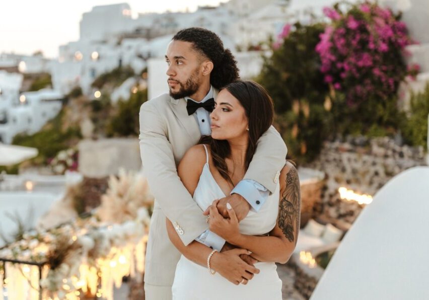 A man in a light suit embraces a woman in a white dress outdoors, with white buildings and pink flowers in the background.