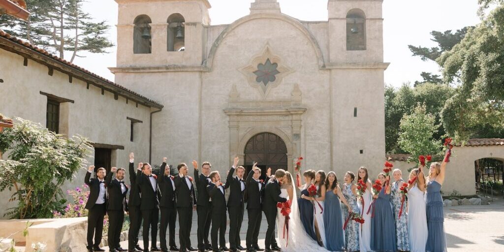A wedding party poses in front of a historic church, with bridesmaids in blue dresses and groomsmen in black suits holding red bouquets and raising their hands.