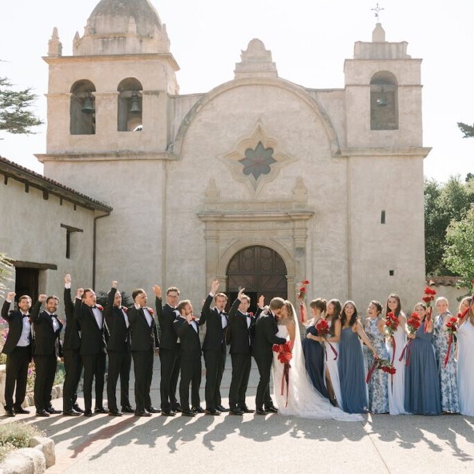 A wedding party poses in front of a historic church, with bridesmaids in blue dresses and groomsmen in black suits holding red bouquets and raising their hands.
