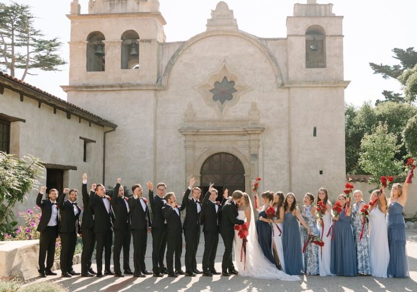 A wedding party poses in front of a historic church, with bridesmaids in blue dresses and groomsmen in black suits holding red bouquets and raising their hands.