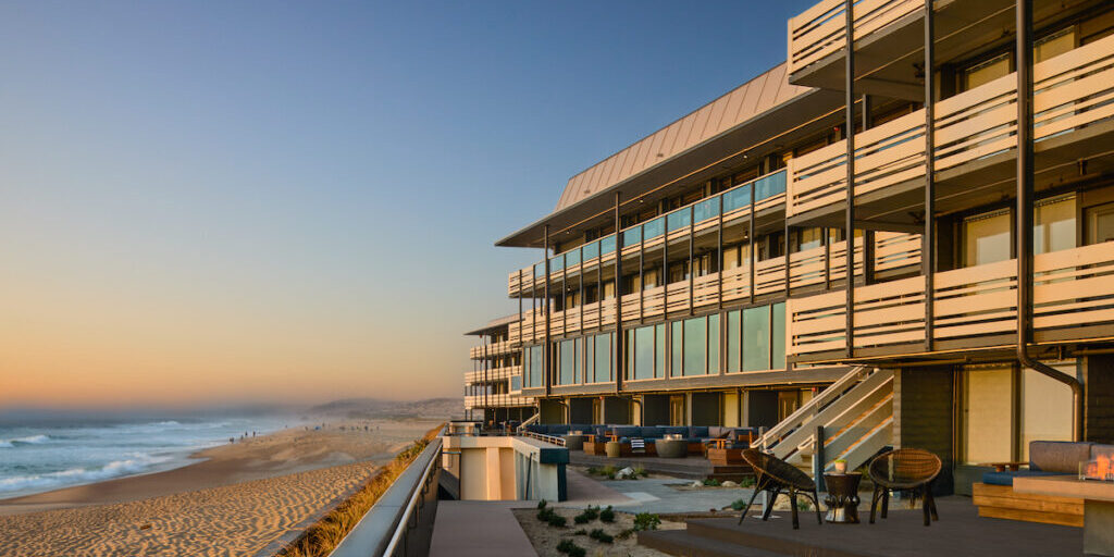 Modern beachfront building with large windows overlooking a sandy beach and ocean at sunset, featuring outdoor seating areas and a boardwalk.