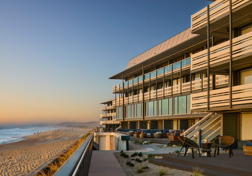 Modern beachfront building with large windows overlooking a sandy beach and ocean at sunset, featuring outdoor seating areas and a boardwalk.