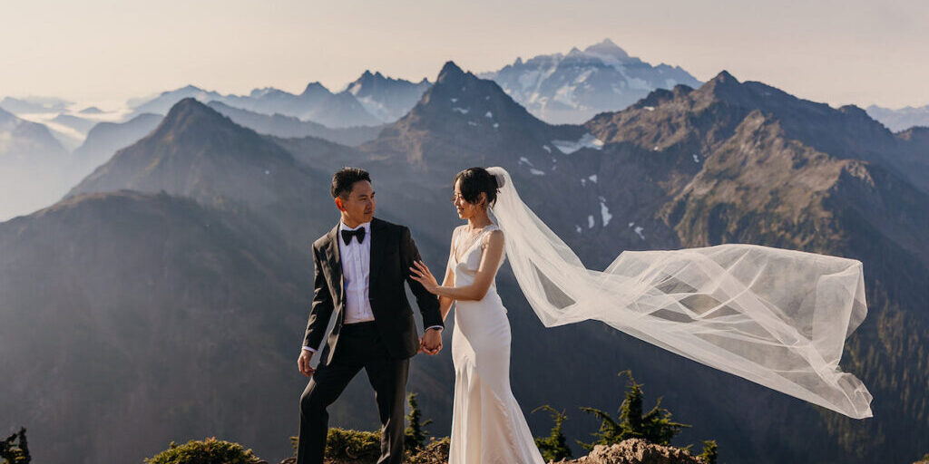 A bride and groom stand on a mountain ledge, holding hands, with mountains in the background and the bride's veil flowing in the wind.