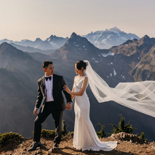 A bride and groom stand on a mountain ledge, holding hands, with mountains in the background and the bride's veil flowing in the wind.