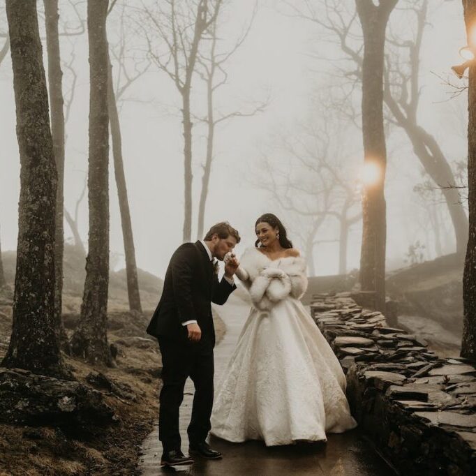 A couple in formal attire walks in a misty forest. The woman wears a white gown and fur stole, while the man dons a suit. They walk along a stone path surrounded by leafless trees.