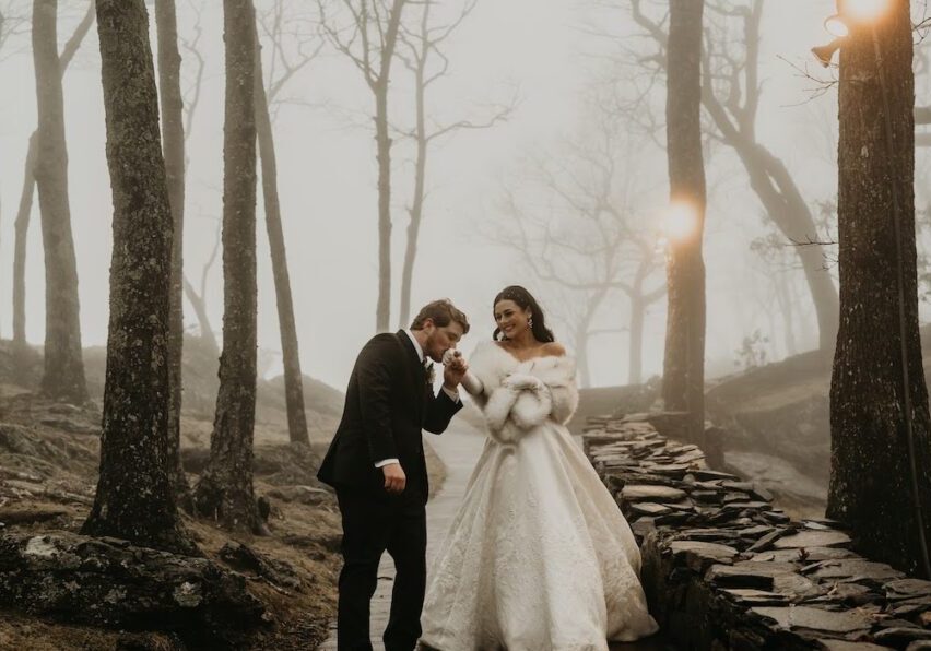 A couple in formal attire walks in a misty forest. The woman wears a white gown and fur stole, while the man dons a suit. They walk along a stone path surrounded by leafless trees.