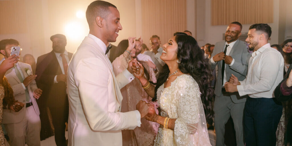 A couple in formal attire dances joyfully at a wedding reception, surrounded by smiling guests on the dance floor.