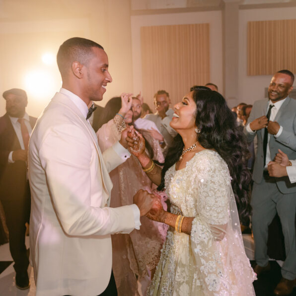 A couple in formal attire dances joyfully at a wedding reception, surrounded by smiling guests on the dance floor.