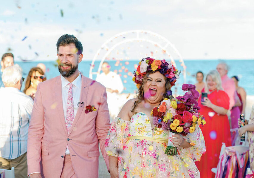 A couple walks down the aisle at an outdoor wedding by the sea, smiling and holding hands, with confetti in the air and guests in the background.