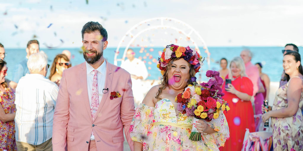 A couple walks down the aisle at an outdoor wedding by the sea, smiling and holding hands, with confetti in the air and guests in the background.
