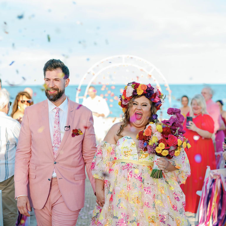 A couple walks down the aisle at an outdoor wedding by the sea, smiling and holding hands, with confetti in the air and guests in the background.