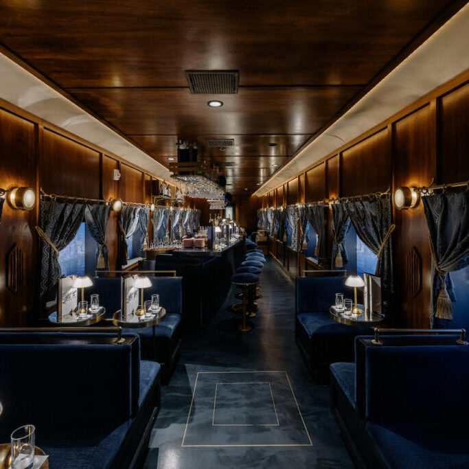 Luxurious train dining car with dark wood paneling, blue upholstered seats, small elegant lamps, and large windows with cityscape views.
