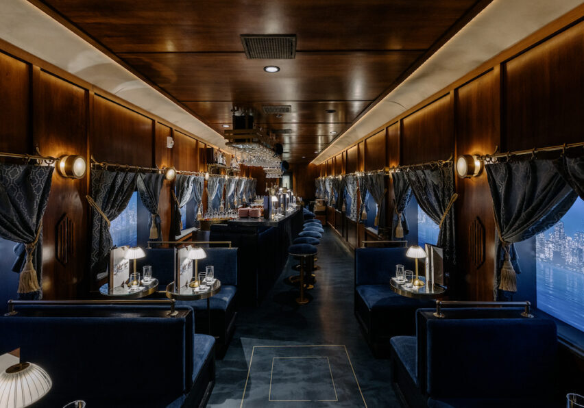 Luxurious train dining car with dark wood paneling, blue upholstered seats, small elegant lamps, and large windows with cityscape views.