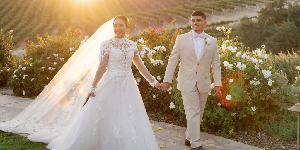 Bride and groom walk hand in hand outside at sunset, with vineyards and white flowers in the background.