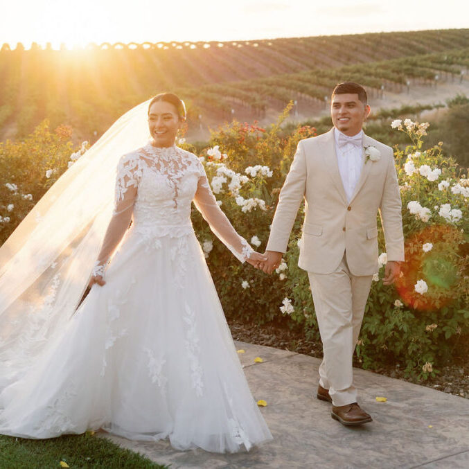 Bride and groom walk hand in hand outside at sunset, with vineyards and white flowers in the background.