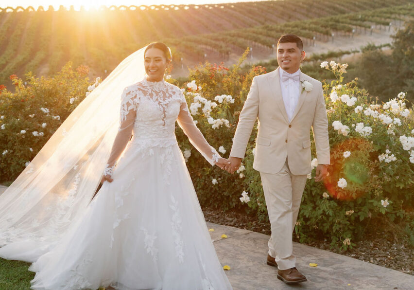 Bride and groom walk hand in hand outside at sunset, with vineyards and white flowers in the background.
