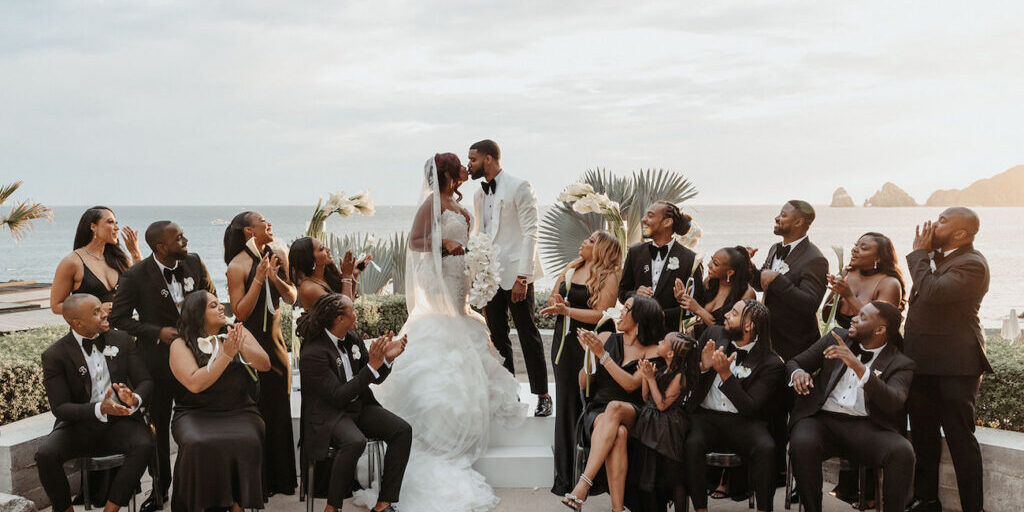 A bride and groom stand at an outdoor altar by the sea, surrounded by a wedding party dressed in black, clapping and smiling.