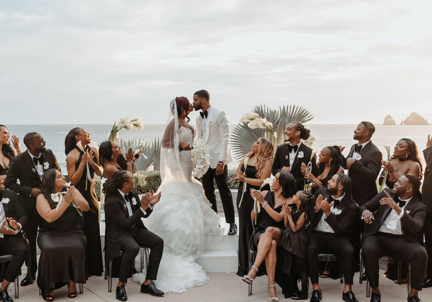 A bride and groom stand at an outdoor altar by the sea, surrounded by a wedding party dressed in black, clapping and smiling.