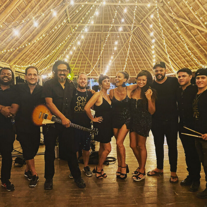 A group of musicians dressed in all black smiling under a thatched roof with string lights
