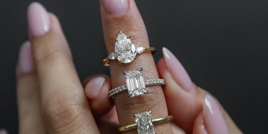 A hand with manicured nails displays three diamond rings of different shapes and bands on the index, middle, and ring fingers against a dark background.