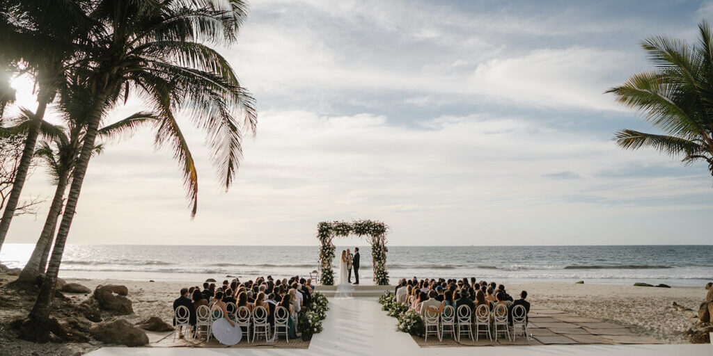 A wedding ceremony takes place on a beach, with guests seated facing an altar decorated with flowers, under palm trees by the ocean.