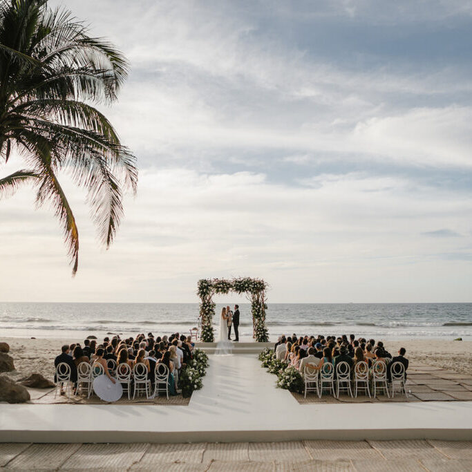 A wedding ceremony takes place on a beach, with guests seated facing an altar decorated with flowers, under palm trees by the ocean.
