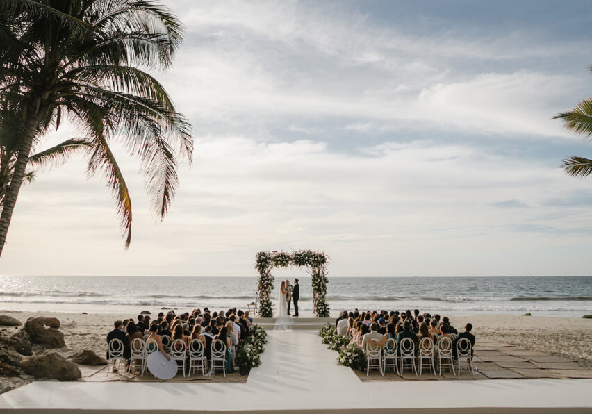 A wedding ceremony takes place on a beach, with guests seated facing an altar decorated with flowers, under palm trees by the ocean.