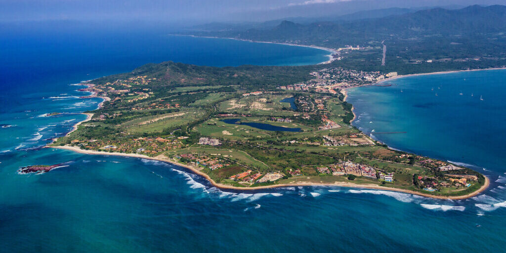 Aerial view of a coastal town with green landscape, buildings, and curving shoreline bordered by blue ocean waves.