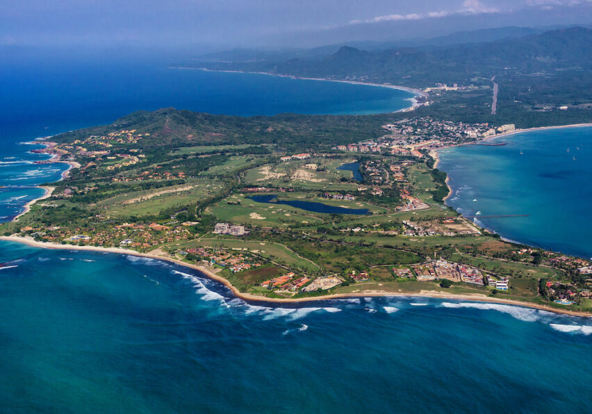 Aerial view of a coastal town with green landscape, buildings, and curving shoreline bordered by blue ocean waves.
