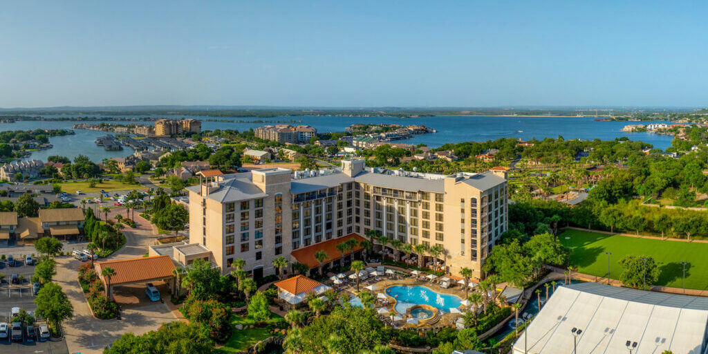 Aerial view of a large hotel with multiple floors, a courtyard swimming pool, palm trees, and a lake with surrounding buildings in the background.