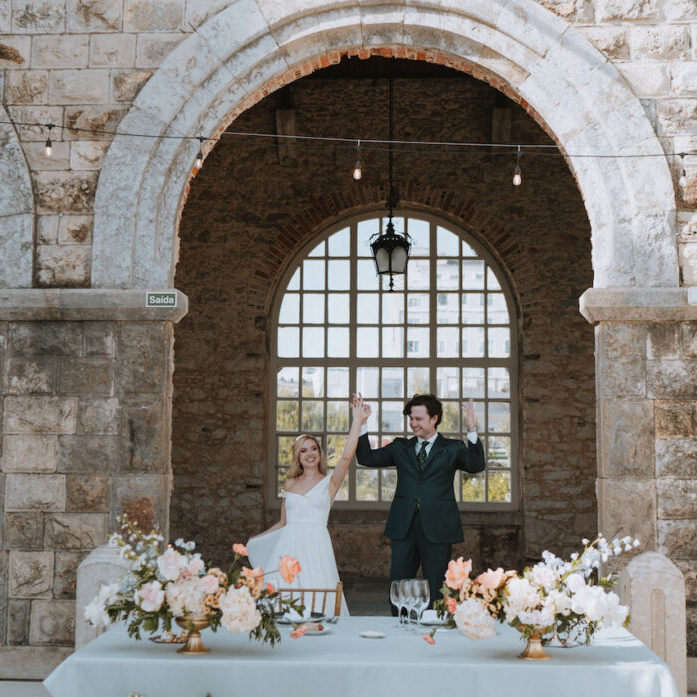 A bride and groom celebrate joyfully at a wedding reception in front of a rustic stone archway, with tables decorated with flowers and glassware.