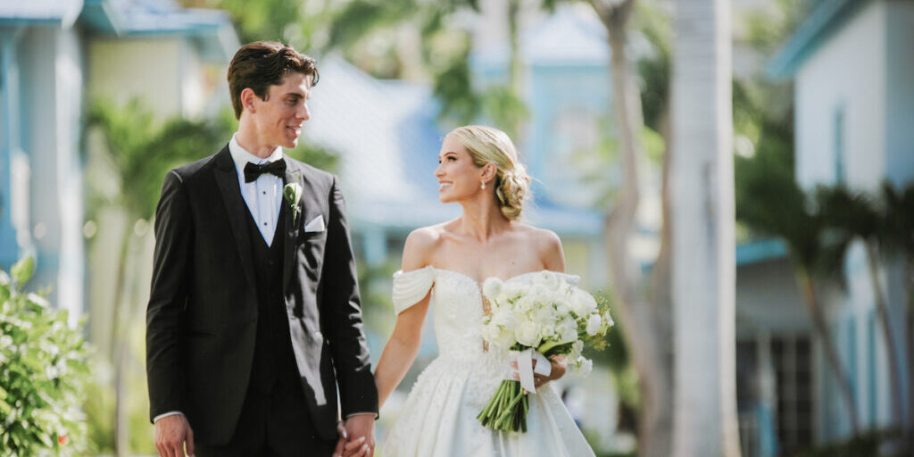 A bride and groom in formal wedding attire hold hands and look at each other while walking outdoors, surrounded by greenery and buildings.