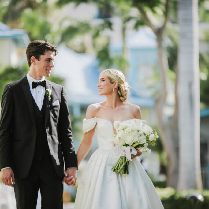 A bride and groom in formal wedding attire hold hands and look at each other while walking outdoors, surrounded by greenery and buildings.