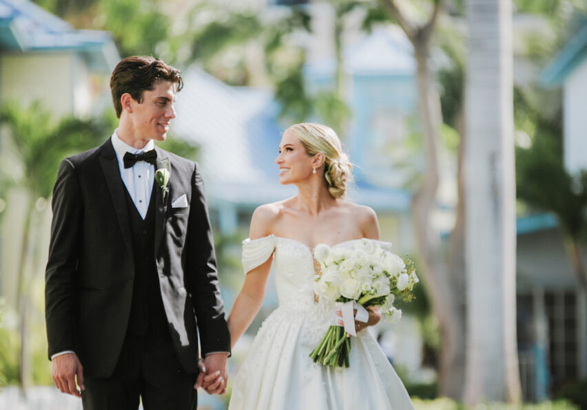 A bride and groom in formal wedding attire hold hands and look at each other while walking outdoors, surrounded by greenery and buildings.