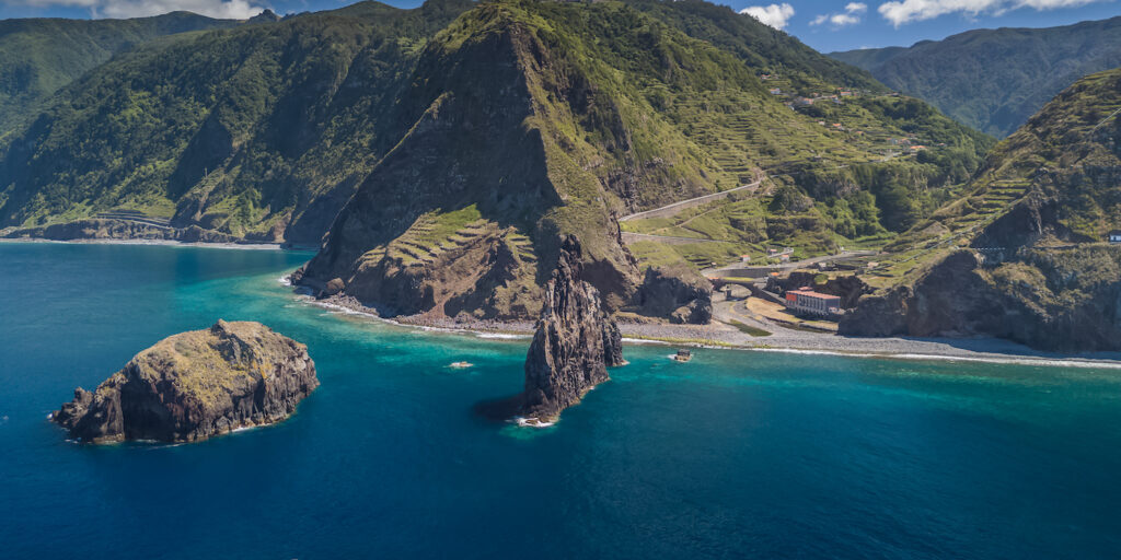 A large rocky peak rises from the turquoise sea near a lush, mountainous coastline with terraced hillsides and a winding coastal road.
