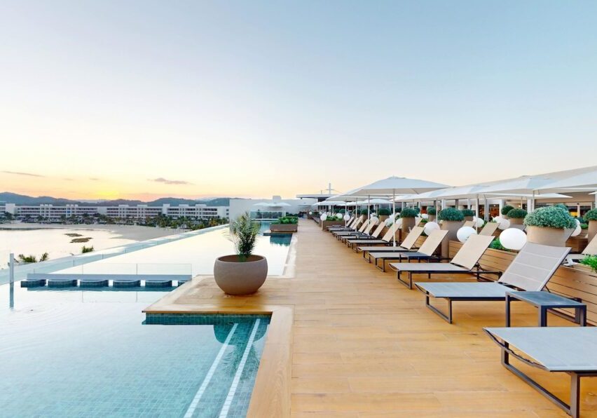 Rooftop infinity pool with lounge chairs, potted plants, and umbrellas at sunset, overlooking a cityscape.