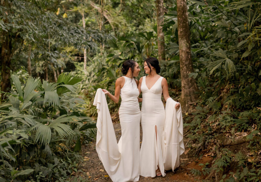 Two brides stand on a forest path, holding hands and gazing at each other, surrounded by lush greenery.