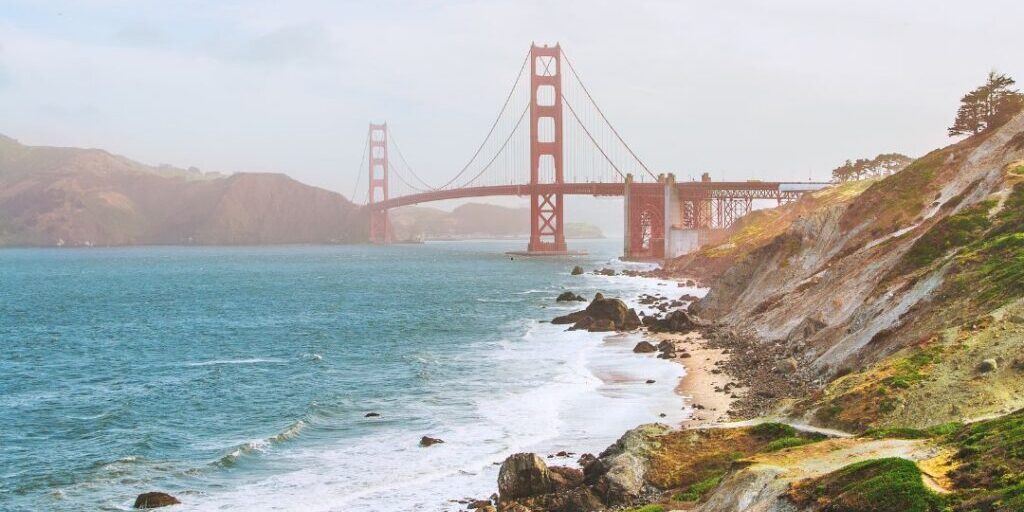 Golden Gate Bridge spans across the water with waves crashing along a rocky shoreline under a cloudy sky.