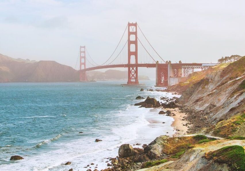 Golden Gate Bridge spans across the water with waves crashing along a rocky shoreline under a cloudy sky.