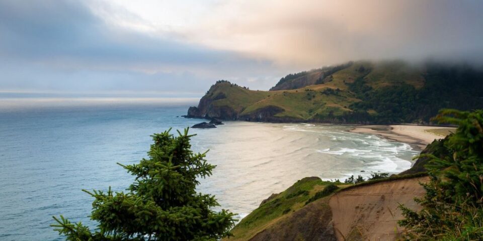 A coastal landscape with a sandy beach, ocean waves, rocky cliffs, and a misty hill under a cloudy sky.