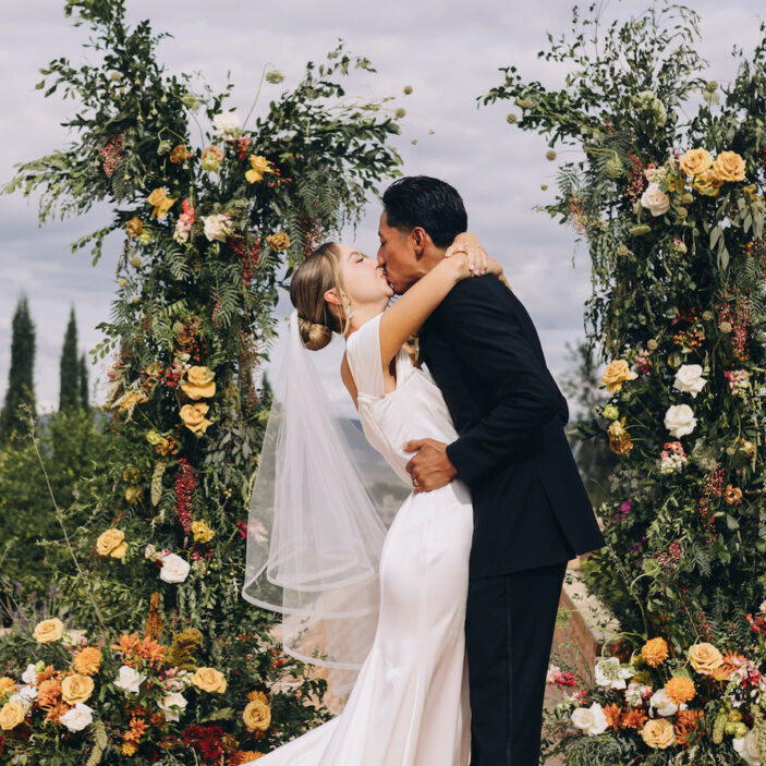 A bride and groom share a kiss at their wedding ceremony, standing between two floral arches, with greenery and mountains in the background.