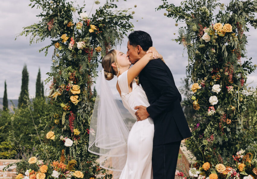 A bride and groom share a kiss at their wedding ceremony, standing between two floral arches, with greenery and mountains in the background.