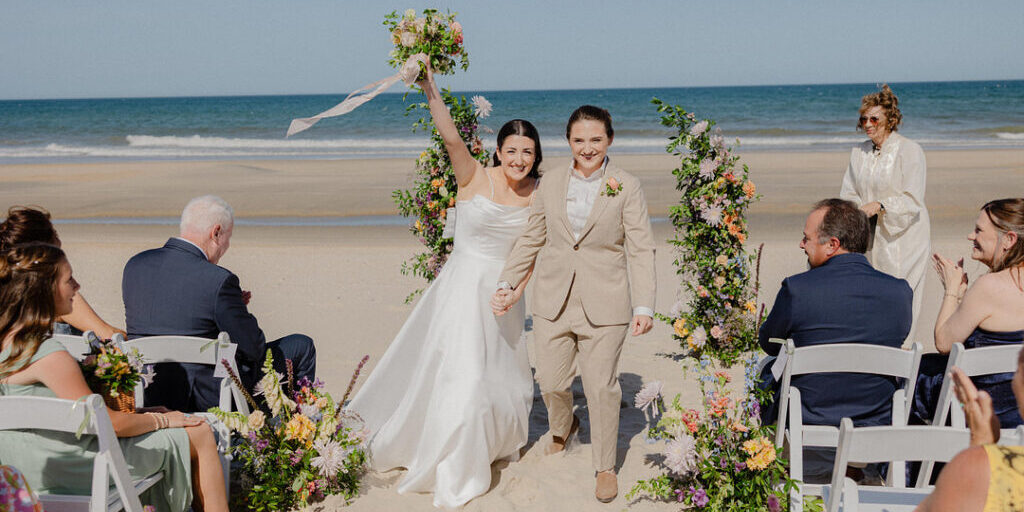 Two women, one in a white dress and one in a tan suit, walk hand-in-hand down a beach aisle lined with flowers, celebrating after their wedding ceremony.