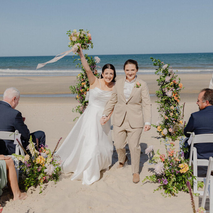 Two women, one in a white dress and one in a tan suit, walk hand-in-hand down a beach aisle lined with flowers, celebrating after their wedding ceremony.