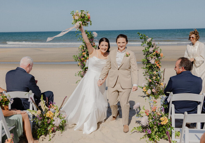 Two women, one in a white dress and one in a tan suit, walk hand-in-hand down a beach aisle lined with flowers, celebrating after their wedding ceremony.