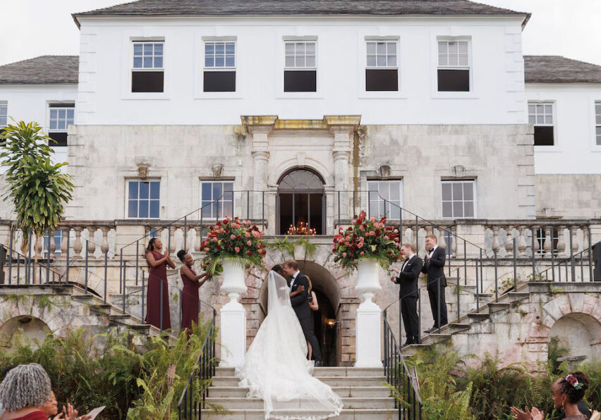 A bride and groom kiss on the steps of a large stone building, surrounded by their wedding party and guests.