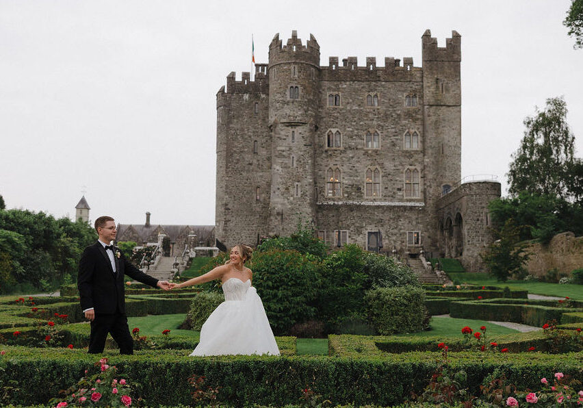 A bride and groom hold hands in a manicured garden with a large stone castle in the background.