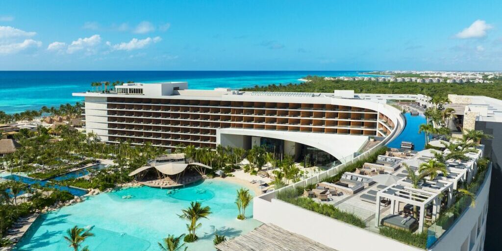 A modern beachfront resort with a curved multi-story building, outdoor pool, palm trees, and views of turquoise ocean under a clear blue sky.