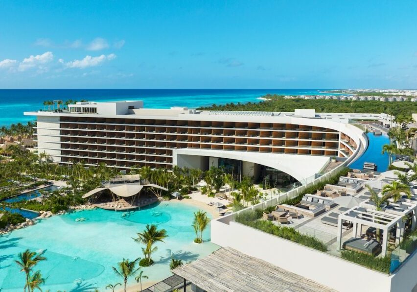A modern beachfront resort with a curved multi-story building, outdoor pool, palm trees, and views of turquoise ocean under a clear blue sky.