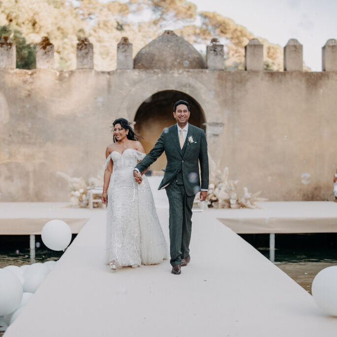 A bride and groom walk hand in hand down an outdoor aisle, smiling, with a stone wall and arches in the background and white balloons along the walkway.